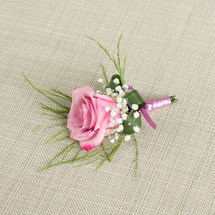 A romantic pink rose boutonniere is beautifully showcased against a pale beige, linen-like fabric that brings to mind the relaxed, natural elegance of homes and venues around Stanmore Hill. The image is taken from a slightly overhead angle, allowing the viewer to see every detail of the compact arrangement. At its centre sits a single rose, its bloom in a semi-open state where the inner petals remain gently cupped while the outer layers curl outward with a graceful, velvety softness. The colour of the rose moves from a subtle blush pink in the heart to a more saturated, slightly deeper rose hue at the petal tips, capturing that fleeting moment of freshness just after the flower has fully awakened. Clustered closely around the base of the rose are multiple stems of delicate white baby's breath, forming a dainty collar of tiny, star-like blossoms that lend the piece an ethereal, almost cloud-like texture. From behind the rose, fine strands of bright green, grass-like foliage radiate outward, introducing movement and lightness, while a broader, darker green leaf is folded artfully to encircle the stems, adding structure and ensuring the boutonniere sits neatly on a lapel. The stems are carefully bound with a shimmery two-toned satin ribbon in lavender-pink and rich magenta, wrapped smoothly and tied in a tidy knot that speaks of skilled florist hands, such as those at Flower Delivery Stanmore who regularly prepare wedding flowers for local ceremonies and celebrations. Soft, diffused light illuminates the scene, catching the sheen of the ribbon, the gentle weave of the fabric, and the subtle shadows between the petals, resulting in a calm, romantic composition perfect for grooms, groomsmen, or prom nights across Stanmore and the surrounding area.
