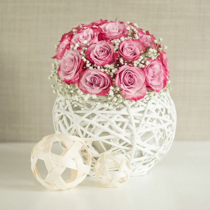 An elegant floral vignette displays a rounded bouquet of pink roses and white baby's breath arranged in a contemporary white woven vase, styled on a pristine white surface against a warm neutral wall, as if set in a tranquil Stanmore home. The bouquet itself is lush and full, composed of numerous roses whose petals show a soft gradient from pale pastel pink at the core to a deeper, almost raspberry pink along the outer edges, giving a gentle ombré effect. Each bloom appears carefully placed, their heads forming a smooth, domed silhouette. Interspersed between these roses are frothy sprays of Gypsophila, with countless tiny white blossoms that create a light, cloud-like texture and soften the rich pinks. The stems are mostly hidden inside a spherical vase constructed from white, interwoven strands that resemble painted wicker or rattan, creating an open basket-weave structure. This airy pattern lets light slip through, forming delicate shadows on the glossy white tabletop below. Set to the left of the vase are two decorative spheres of different sizes made from thin, natural wood or rattan strips in a pale beige tone. Their open, geometric design mirrors the lattice of the vase, tying the scene together through repeating shapes and textures. Behind everything, a softly textured beige or cream wall provides a calm, unobtrusive backdrop, much like the soothing interiors found in modern apartments near Stanmore station or family homes off Marsh Lane. The overall atmosphere is one of fresh, modern romance with a hint of rustic charm, suitable for a styled coffee table, an anniversary arrangement, or a thoughtful gift arriving via local delivery. The image reflects the kind of carefully composed design a professional florist in Stanmore would prepare for special moments.