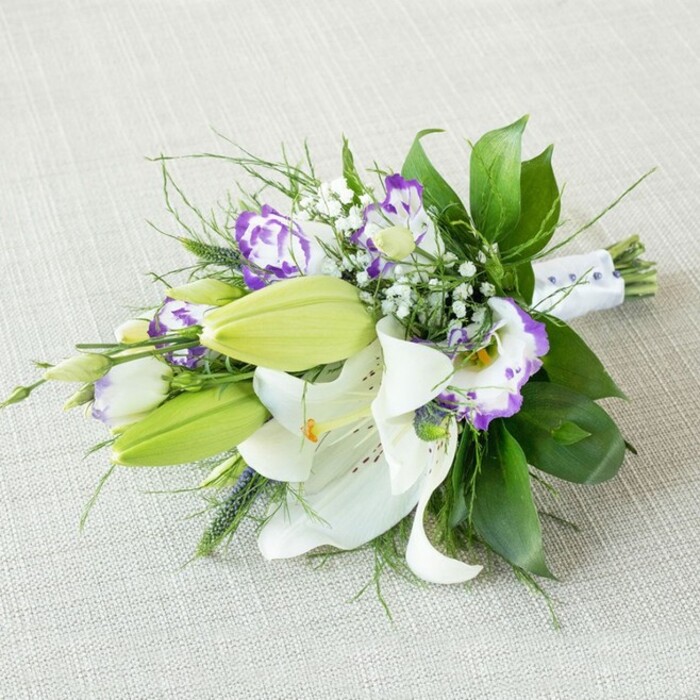 An elegant, elongated bridal-style bouquet is arranged diagonally across a light beige, subtly textured fabric, the simple background echoing the calm, understated charm often associated with homes around Stanmore. The composition is dominated by generous white Oriental lilies, several of them fully open so that their broad petals sweep outward in smooth, graceful arcs. Each lily reveals bold yellow-orange stamens dusted with pollen, standing in contrast to the pale, almost luminous petals, which are delicately speckled with fine reddish-brown markings close to the center. Between these open blooms, a number of fresh, light green lily buds rise on firm stems, their tightly closed forms suggesting the bouquet will continue to change and bloom after it leaves the florist's bench. Interspersed among the lilies are clusters of Lisianthus (Eustoma) blossoms, their ruffled petals forming layered cups of white kissed with vivid purple and violet edges, the colour softly deepening toward the centers like watercolour washes. Fine sprays of white Gypsophila thread through the bouquet, the tiny, cloud-like flowers breaking up the larger shapes and lending an airy, romantic softness. A lush foundation of greenery supports the design: broad, glossy dark green leaves create a strong visual base, while slender, wispy fronds and a few lightly spiked stems add movement and texture, reminiscent of foliage found in nearby Bentley Priory Nature Reserve. The stems are gathered tightly at one end and wrapped in a crisp white satin ribbon, finished with small, pearl-like beads or delicate purple accents that hint at bridal detailing without overwhelming the flowers. Gentle, diffused lighting washes over the arrangement, emphasizing the creamy whites, fresh greens, and rich purples while avoiding harsh shadows, creating a serene, almost timeless atmosphere. The result is a bouquet that radiates purity, romance, and quiet sophistication, perfectly suited to a Stanmore wedding, an elegant engagement celebration, or a thoughtful floral gift ordered through Flower Delivery Stanmore for local delivery.