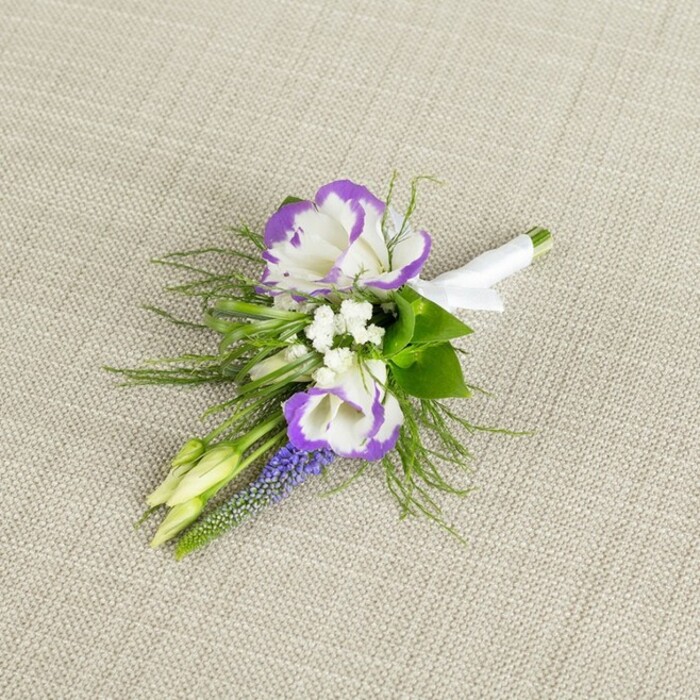 An elegant boutonniere is displayed against a softly textured, light beige linen background, the warm neutral tone quietly framing the floral details without distraction. At the centre, two prominent lisianthus blossoms unfurl their white petals, each delicately ruffled and edged with a rich, royal-purple border that creates a striking contrast and a sense of refined drama. Around them, tiny white filler flowers-similar to baby's breath-gather in subtle clusters, their miniature blooms lending a lace-like softness that tempers the boldness of the purple edges. A slender stem of muscari, or grape hyacinth, runs vertically through the design, its tightly packed, bell-shaped flowers coloured in a deep, grape-toned violet that echoes the lisianthus edges while introducing a new, textured rhythm. Fine, feathery greenery arcs gracefully around the blooms, bringing a light, airy frame of fresh green that suggests something handpicked from a shaded corner near one of Stanmore's quiet residential gardens or from the paths around Stanmore Country Park. Two pale green buds remain unopened within the arrangement, adding a sense of anticipation and new beginnings, fitting for a wedding morning or prom evening. The stems are carefully aligned and encased in a band of crisp white satin ribbon, smoothly wrapped and neatly tied, indicating expert florist craftsmanship and making it easy to pin to a lapel. The soft, even lighting highlights the interplay of white, purple, and green, creating a calm, sophisticated mood. This small yet expressive piece feels ideal for a groom or groomsman preparing for a ceremony in Stanmore, carrying the gentle fragrance and quiet emotion of a thoughtfully chosen floral detail close to the heart.
