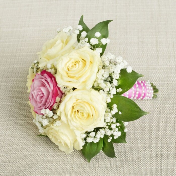 This charming bouquet, captured from an inviting, slightly elevated perspective, exemplifies the refined floral artistry embraced by Stanmore's local flower designers. Featuring an ensemble of soft, creamy-white roses whose petals gently unfurl like morning blooms over Canons Park, the bouquet's palette is accented by a single, striking magenta-pink rose resting near the center, serving as a vibrant signature flourish. An abundance of delicate white baby's breath threads through the collection, casting an ethereal, almost whimsical haze around the roses, while deep green leaves peek from beneath, grounding the bouquet with natural richness. The visible handle, wrapped in a vivid fuchsia-pink satin ribbon, showcases polished elegance and is adorned by a spiraling string of white pearls, sparkling softly in ambient light. Resting upon a beige, textured linen reminiscent of welcoming Stanmore interiors, the arrangement's colours and textures are celebrated in every detail. This bouquet is a sophisticated choice for brides, bridesmaids, or celebrants seeking effortless grace at local venues, and reflects the pride taken in every creation by the Flower Delivery Stanmore team, renowned for their devotion to quality and heartfelt connection to the community.