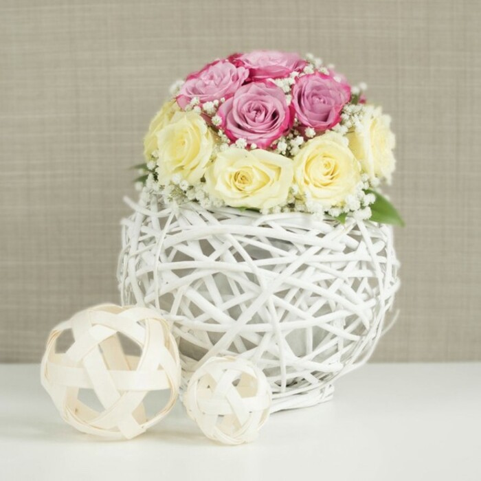 A meticulously composed floral display is shown in close-up, evoking the quiet elegance of a wedding table set in Stanmore's leafy neighbourhoods. The focal point is a rounded bouquet made up of around a dozen fresh roses, carefully arranged so their heads form a smooth, domed silhouette. Soft pastel pink blooms with a subtle lavender or fuchsia undertone mingle with creamy yellow and ivory roses, the interplay of hues creating a gentle contrast that feels both romantic and timeless. Each rose tells a slightly different story: some are still half-closed, their centers hidden deep within tightly wrapped petals, while others are more open, revealing intricate spirals and layered folds that suggest fullness and luxury. Fine, branching stems of baby's breath thread among the roses, their tiny white blossoms generating a misty, cloud-like haze around the flowers and giving the arrangement an airy, delicate finish. Cradling the bouquet is a distinctive spherical vase made from slim white branches or wicker, woven into a complex open lattice that resembles a decorative globe. The open-weave structure allows hints of the inner container to peek through, a subtle detail that showcases the florist's structural work. This decorative sphere rests on a crisp white tabletop, smooth and unadorned, drawing attention to the floral artistry. To the left sit two smaller decorative orbs constructed from light, natural wood strips in a similar open pattern, one larger and one smaller, reinforcing the cohesive, textural theme. The background is a neutral beige or light taupe with a faint fabric-like texture, similar to the soft interiors of homes near Stanmore Country Park or along Old Church Lane. Altogether, the scene radiates calm sophistication and gentle romance, perfect for a wedding breakfast, anniversary celebration, or intimate dinner styled by a local Stanmore florist.