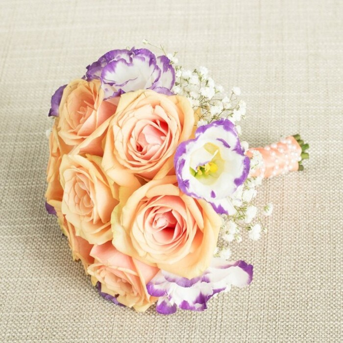 This thoughtfully arranged bouquet sits poised on a subtle, linen-textured background, evoking the tranquil interiors and graceful gatherings of Stanmore's elegant homes. The arrangement's warm foundation is formed by large, full peach and apricot roses-each blossom unfurling creamy petals tinged with blush pink, suggesting softness and luxury. Mixed among these are striking lisianthus blooms, pure white and vibrantly rimmed with purple, a specialty bloom often admired in Stanmore's local florists. Clouds of delicate white baby's breath nestle in between, lending the whole bouquet a breath of airy volume, reminiscent of Stanmore Country Park's open spaces. The stems are wrapped in shimmering peach satin ribbon and finished with subtle pearl beads-a handcrafted detail that brings a timeless and polished feel, suited perfectly to weddings and landmark occasions locally. The bouquet's round, collected form feels as if it was made for cherished moments: held tightly during vows in a Stanmore church, or gracing a table at a family celebration. With its harmonious blend of pastel hues and precisely selected accents, this bouquet captures both the romantic spirit and sophisticated taste unique to the Stanmore area.