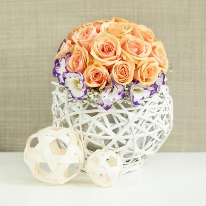 Set against a softly textured beige backdrop that hints at linen or fine-woven fabric, this floral centerpiece brings a sense of calm celebration, as though prepared for an intimate gathering in a Stanmore living room near the high street. Rising from the centre is a perfectly rounded dome of roses in peach, apricot, and gentle salmon hues. The blooms are closely packed, their petals fully unfurled and overlapping, creating a smooth, almost sculpted form with subtle colour gradations that move from pale, creamy edges to deeper, glowing hearts. Surrounding the lower edge of this rose dome is a band of white lisianthus, each bloom edged with a distinct strip of deep violet and purple. The frilled, slightly curling petals add movement and intricacy, while fine sprays of gypsophila slip between them, their tiny white flowers softening the contrast and lending a whisper-light texture. All of the flowers are gathered into a distinctive, broadly spherical container crafted from white rattan or wicker strands. The open lattice design of the vase forms a bulbous, nest-like sphere, allowing light and shadow to intermingle and giving the piece an airy, contemporary feel that still nods to natural materials. On the white surface before the arrangement are two decorative orbs woven from pale beige, wood-like strips. One orb is larger, the other smaller, and both repeat the open, intertwined motif of the main container, visually tying the elements together. The overall effect is sophisticated yet approachable, ideal for illustrating wedding centrepieces, engagement party décor, or elegant table flowers available for same-day delivery across Stanmore. The arrangement captures the gentle luxury and relaxed refinement that suit the area's leafy streets and spacious homes.