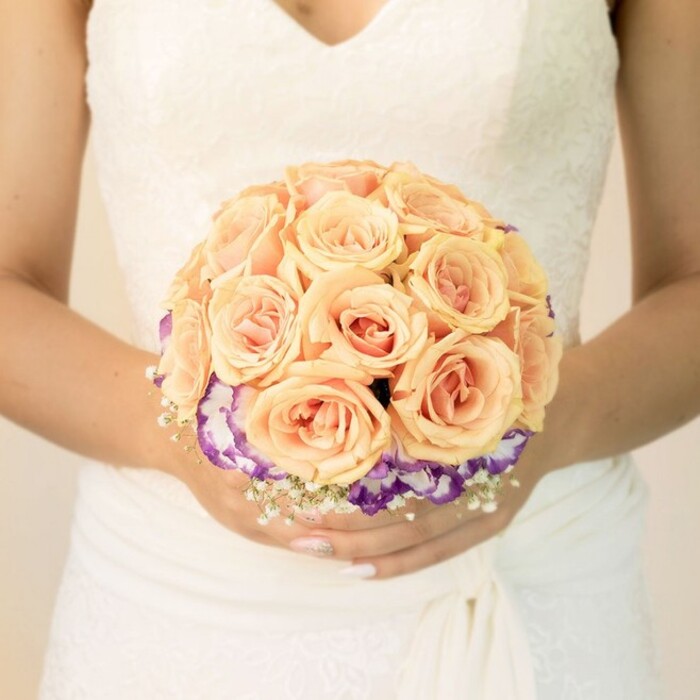 The photograph captures a close, chest-level view of a bride holding her bouquet, the background blurred to a soft, neutral wash that could easily be the light walls of a Stanmore town hall or a sunlit room overlooking Bentley Priory Nature Reserve. She is dressed in a traditional white or ivory gown, the bodice delicately worked with lace that draws the eye upward to a modest V-neckline. The fabric at her waist is gently ruched or wrapped, creating a flattering, sculpted effect, while the skirt flows downwards in smooth, unbroken lines just visible beneath her hands. At the centre of the frame sits her bridal bouquet, crafted with the care of an experienced florist who understands both structure and sentiment. The bouquet is round and compact, dominated by a generous cluster of roses in soft peach, apricot, and blush pink hues. Each rose appears freshly opened, petals tightly layered and curling inwards to form lush, velvety spirals, the colours blending from pale outer edges into warmer, deeper centres. Beneath this rounded crown of roses, a vivid ring of deep purple flowers with white, ruffled margins-likely lisianthus or a similar bloom-circles the base, adding dramatic contrast and visual depth. Intertwined among these are wisps of tiny white flowers, reminiscent of baby's breath, which soften the outline and create a light, cloudlike fringe. The bride's hands cradle the stems with quiet assurance; her nails are short and neatly manicured in a light, natural shade, with a touch of shimmer on one thumb that hints at her personal style. The overall mood is serene and romantic, ideal for showcasing how Stanmore wedding floristry, including designs prepared by local specialists such as Flower Delivery Stanmore, can blend gentle colour transitions with thoughtful textural details to create unforgettable, photograph-ready bouquets.