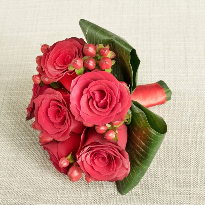 A sophisticated hand-tied cluster of red roses and berries is displayed against a pale beige, subtly textured fabric, evoking the calm of a softly lit Stanmore bedroom or sitting room. Around six full-bodied roses take centre stage, each blossom boasting layers of velvety petals that shift from dark ruby at the core to slightly brighter crimson along the outer edges. Their rounded shapes nestle closely together, creating a compact, almost sculptural dome of colour. Nestled between the rose heads, tight clusters of small, round Hypericum berries in deep red and reddish-pink introduce a lively, tactile contrast, like tiny polished beads sprinkled throughout the bouquet. Embracing the arrangement are broad, dark green leaves, possibly aspidistra or a similar tropical foliage, carefully folded and curved with an experienced florist's touch to wrap partially around the blooms. This green collar adds a sense of depth and a sleek, modern line that makes the red tones appear even richer. At the base, a band of red satin ribbon is wrapped neatly around the stems, its smooth, reflective surface hinting at fine gift-wrapping and thoughtful presentation. Gentle, even lighting casts soft highlights across the petals, berries, and foliage, with no harsh shadows, allowing every detail to be clearly appreciated. The bouquet feels ideal as a romantic gesture sent to a loved one near Stanmore station, or as a refined centrepiece for an intimate dinner in a home off The Broadway. It conveys classic passion with a contemporary twist, the kind of carefully crafted design you might expect from a skilled local florist who understands how Stanmore families celebrate life's important moments.