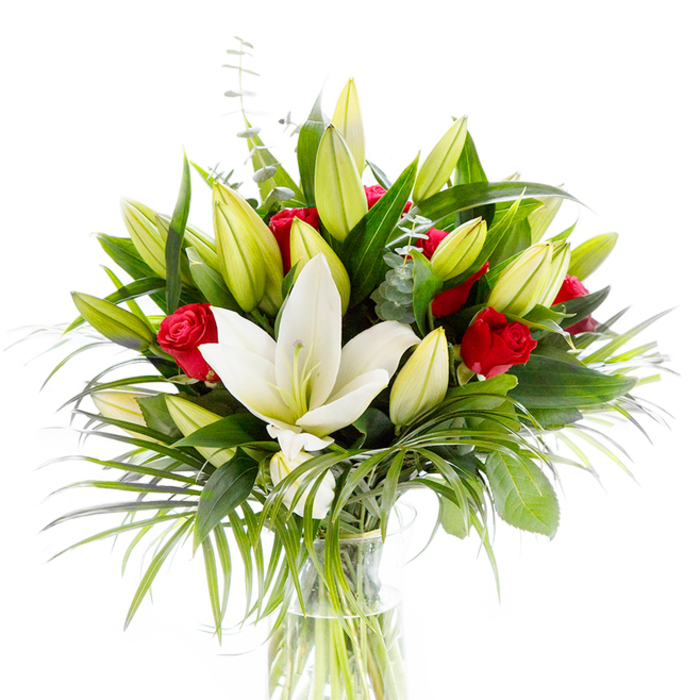At the centre of this composition sits a majestic white Oriental lily, wide open and luminous against a bright white backdrop, its smooth cream petals cupping a delicate yellow stamen and pale green pistil like a small, serene heart. Around it, clusters of lily buds in lime and soft yellow hues create architectural volume - tight, rounded buds that promise new blooms and bring a gentle progression of form. Interspersed among the lighter lilies are compact red rosebuds and small roses, their velvety petals still furled into intimate spirals that lend a classic, passionate accent to the otherwise pale palette. The foliage is abundant and varied: deep, lustrous forest-green leaves provide a strong framework; long, feathered palm fronds in cleaner emerald tones cascade softly away from the centre, adding drama and a whisper of the exotic; and slim eucalyptus stems with their silvery-green, rounded leaves weave through the arrangement and soften the overall effect. The flowers are set in a plain, cylindrical clear glass vase, the kind a florist favours for transparency and honesty - the pale green stems are visible submerged in clear water, and the studio's bright white lighting makes every edge, curve, and texture read clearly. Visually and olfactorily the bouquet balances purity and passion: the lilies' clean, powdery scent lifts against the roses' deeper, sweeter perfume. This arrangement feels at once formal and tender, suitable as a wedding gesture, anniversary tribute, or centrepiece for a celebratory table, and it carries the quiet domestic familiarity of bouquets often selected by households around Stanmore Broadway and the green walks of Stanmore Common.
