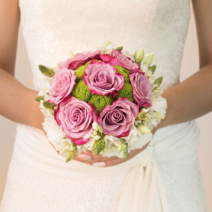 A moment of bridal serenity and artistry is captured in this close-up, where a bouquet of richly coloured blooms is gently held against the bride's flowing lace gown-a scene that could unfold at any cherished Stanmore wedding venue. At the centre, velvety roses with petals graded from a gentle lavender to vivid dusty pink, their outlines touched with vibrant fuchsia, command attention and evoke a sense of deep romance. Crowded around these central roses are brightly toned lime-green button chrysanthemums, their compact, textured heads providing a cheerful contrast suggestive of the well-kept lawns found along Stanmore's leafy roads. The boundary of the bouquet is softened by tufts of delicate white flowers-perhaps clusters of freesia or miniature spray roses-contributing to an air of purity and gentle movement. Small buds and slender greens thread through the outer ring, enhancing both the sense of freshness and compositional depth. The bride's hands, nails accented in soft pink with a subtle shimmer, embrace the stems with care, while the patterned lace of her bodice reflects the understated elegance typical of Stanmore's community. The play of natural light reveals each petal, cluster, and subtle shade, framing the entire arrangement in a tranquil, joyful ambiance. Lovingly composed by a Stanmore florist, this bouquet celebrates not only the couple's love but the beauty and spirit of this North West London neighbourhood.
