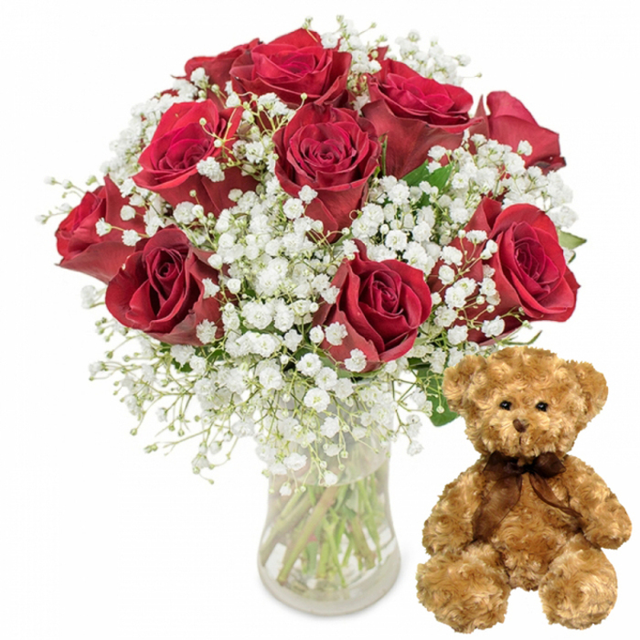 The image captures a classic romantic ensemble displayed as though it has just been arranged in a Stanmore florist's studio near the high street. On the left, a rounded bouquet of deep crimson red roses rises from a clear glass vase. There are around twelve roses, each one with thick, velvety petals that layer together in perfectly formed blooms. The roses show subtle variations in stage: some are luxuriantly open, showing off their inner spirals, while others remain slightly tighter, lending a sense of freshness and life. Weaving gently among the red blooms, sprays of white baby's breath (Gypsophila) form a cloud-like frame. Their clusters of tiny white flowers add a light, almost ethereal contrast that softens the intensity of the roses, giving the arrangement a dreamy quality. Through the transparent glass of the vase, straight green stems converge into clear water, with glimpses of leaves and natural texture providing a grounded, organic feel. To the right of the vase sits a charming teddy bear, about the height of the floral display. Its soft, light brown plush fur looks touchably warm, with a slightly tousled surface that suggests comfort and coziness. The bear's small black button eyes and rounded nose are neatly stitched, creating a gentle expression that seems to smile softly. Around its neck, a slim dark brown satin ribbon is tied into a tidy bow, adding a hint of elegance to its cuddly form. The pure white background and even, shadow-free lighting draw the viewer's focus entirely to this pairing of roses and teddy bear, a scene that captures the sentiment behind many local Stanmore flower deliveries-thoughtful gestures for Valentine's Day, wedding anniversaries, or heartfelt surprises arriving at homes near Stanmore Country Park.