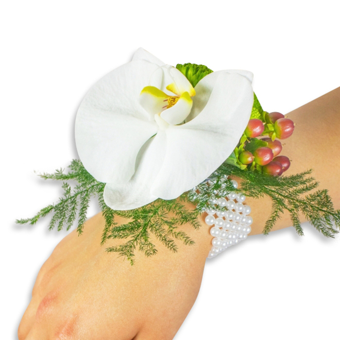 A single, immaculate white Phalaenopsis orchid forms the heart of this elegant wrist corsage, its wide, velvety petals unfurling to reveal a vibrant yellow lip streaked with deeper orange tones that focus attention and add warmth. The orchid is set against a generous bed of bright asparagus fern, whose soft, feathery texture offers contrast and lifts the bloom visually from the wrist. To the upper-right, a cluster of glossy Hypericum berries provides seasonal colour - a gentle progression from pale green to blush and finally to a deep cranberry red - while a sprig of ruffled lime-green accent foliage introduces another tactile layer. The arrangement is secured to a timeless bracelet of multiple white pearl strands that wrap comfortably around fair skin; the pearls' cool sheen and classic silhouette lend a refined, heirloom quality to the design. Photographed on a clean white background with soft, diffused lighting, the piece reveals careful floral mechanics and an attention to proportion typical of experienced local designers. As a Stanmore-based florist serving clients around Stanmore Broadway, Canons Park and the surrounding Harrow neighbourhoods, Flower Delivery Stanmore often recommends this corsage for bridal parties, proms, and formal gatherings - it reads as both romantic and perfectly composed, a small wearable statement that complements both contemporary and traditional outfits and travels well to local venues and intimate celebrations.