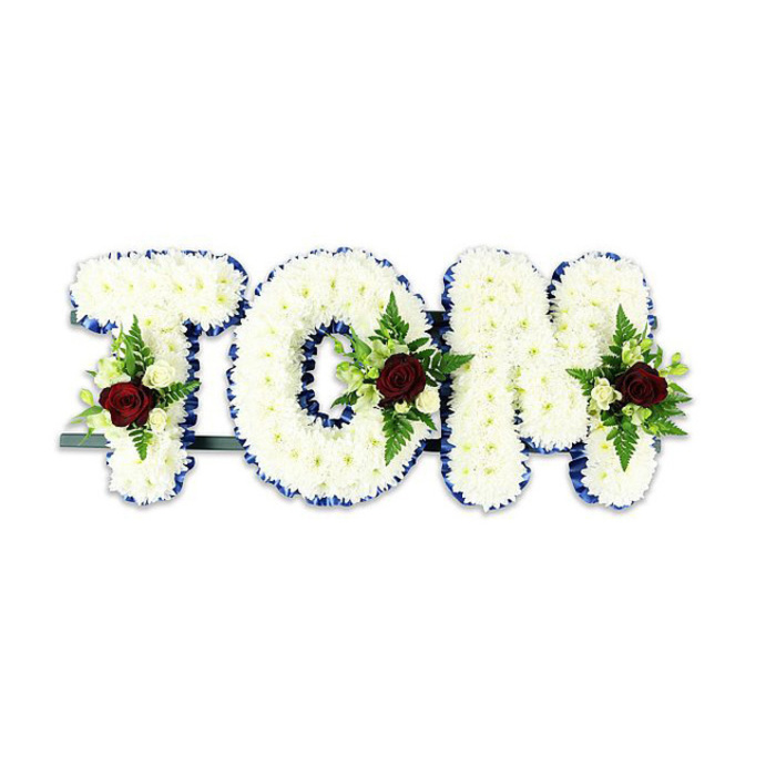 The name TOM is rendered in three-dimensional floral lettering, each letter a compact mosaic of small white chrysanthemum blooms that create a soft, cloud-like surface. Along the outer edges, a narrow royal blue ribbon frames each character, providing a refined contrast that sharpens the silhouette and catches the eye from across a chapel or reception room. Nestled into the centre or base of each letter is a modest spray composed around a single deep red rose; its velvet petals and somber tone immediately draw attention, supported by tiny creamy white spray roses and slender fern fronds that weave between the chrysanthemum heads. The greens introduce a quiet texture - serrated, feathery and alive - while the white chrysanthemums offer a matte, gentle sheen. A dark green or black supporting bar is visible behind the base, indicating the tribute is intended to stand upright during viewings or interment ceremonies. The composition is thoughtful and restrained, designed to be read both as a name and as an emblem of remembrance. As a locally made tribute this style is common for services near Stanmore Broadway or small parish gatherings around Canons Park, where families ask for clear, dignified lettering that reads well in soft daylight and under chapel lighting. The overall impression is one of calm dignity: tactile, uncomplicated, and made to honour a life with measured grace.