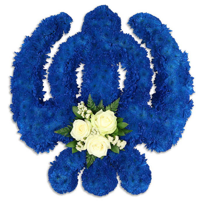 A large, reverent Sikh Khanda tribute dominates the image, presented against a plain white background so that every detail of the floral work can be clearly appreciated. The iconic shape - with its central double-edged sword, its circular chakkar, and its two outstretched kirpans - is carefully defined using a mass of tightly packed royal blue chrysanthemums or similar small, rounded blooms. These flowers form a thick, even surface across the entire symbol, their petals creating a soft, plush texture reminiscent of velvet. The blue is intense yet soothing, suggesting devotion, courage, and spiritual depth, sentiments that resonate strongly with families gathering in Stanmore for funeral services or remembrance prayers. At the base of the central blade, where the eye naturally settles, a smaller grouping of white flowers provides a point of calm contrast. Three white roses, unblemished and perfectly formed, are nestled together, framed by light sprays of baby's breath and rich green leaves that lend dimension and a touch of natural wildness. This central cluster appears almost like a blessing at the heart of the emblem, its white tones reflecting purity and peaceful farewell. Gentle, diffused lighting enhances the floral textures and casts faint shadows, giving the Khanda tribute a three-dimensional presence as though it is ready to be carried into a service at a local venue near Stanmore Station or along Church Road. The arrangement feels carefully considered and deeply respectful, a meaningful way for families to express sympathy and honour a loved one's Sikh heritage through fresh, beautifully arranged flowers.
