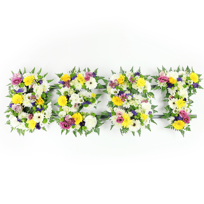Centered against a bright white backdrop, four bold letters spell BEST, each letter lovingly formed from a dense layer of fresh flowers and rich greenery, the kind of tribute we often craft for families across Stanmore and nearby Canons Park. The dominant palette is clear and uplifting: pristine white chrysanthemum-like blooms and cream gerberas create a soft, luminous base; cheerful yellow roses and yellow gerberas add warmth and a sunlit optimism; while lavender-to-purple lisianthus and small purple chrysanthemums bring a hush of dignity. Splashes of vibrant magenta-pink spray roses or carnations provide lively contrast, and clusters of pale green and white filler flowers add fine texture and visual depth. Lush dark green fern fronds trace the outer edges of each letter, giving a gentle, organic border that highlights the handcrafted shapes. Through some tighter areas the dark green metal or subtle support bars peek through - most noticeable along the E and T - showing the careful engineering used to keep large-format letters secure and perfectly formed for display. Shot directly from the front with bright, even light, the composition emphasizes freshness, petal detail, and the soft shadowing that gives each letter dimension. This piece reads as an elegant memorial or celebratory statement, evoking tenderness, respect and joyful remembrance. Prepared with the careful touch of a local florist, the arrangement carries a quiet fragrance of rose and green foliage that seems to echo the peaceful air around Stanmore High Street and the surrounding neighbourhood.