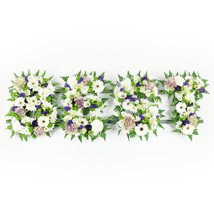 An overhead view presents a horizontal, four-part floral composition spelling the uppercase word BEST, each letter shaped by a dense, sympathetic mix of white and purple blooms set against a pristine white background. Large, pure white Gerbera daisies-with their distinct dark centers-sit as luminous focal points, balanced by clusters of white roses, fluffy carnations and delicate white lisianthus or chrysanthemums, which together create layered textures from smooth petals to rounded fullness. Soft mauve and lavender roses, along with ruffled purple carnations and tiny deeper violet fillers, thread through the letters and add a gentle, dignified shading that suggests warmth and remembrance. Feathery green fern fronds outline and encircle each letter, their soft, natural borders lending a fuller silhouette and a sense of calm motion, while compact green button chrysanthemums punctuate the interior with fresh, round touches of verdant colour. The arrangement rests on a subtle dark grey or black support that occasionally shows through, providing crisp edges and structural clarity to the lettering without competing with the floral surfaces.
The visual tone is reverent and graceful, as suited to a memorial or funeral setting where clarity of message matters as much as the beauty of the blooms. I picture this piece displayed near a service in the Stanmore area-perhaps at a local chapel or alongside tributes on Stanmore High Street-its soft scent of white florals and green foliage offering a quiet, comforting presence. The craftsmanship reveals careful selection and placement, a personalised floral statement that honours the life being remembered with tenderness and composure.