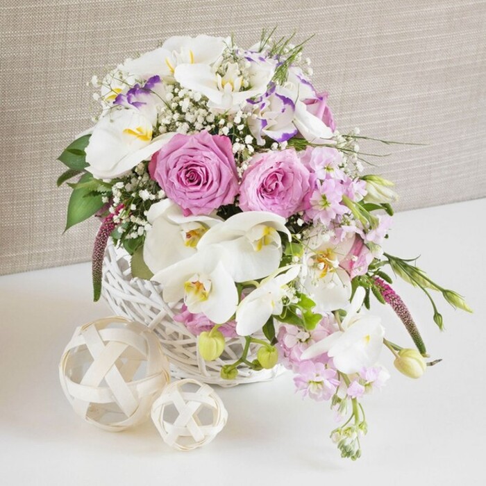 A beautifully composed floral arrangement in a white wicker basket creates a romantic focal point, reminiscent of a wedding table set in one of Stanmore's elegant venues. The basket is round and tightly woven, painted a clean, soft white that frames an abundant collection of pink and white blossoms. At the heart of this design are lush pastel pink roses, each bloom fully opened to reveal complex spirals of velvety petals that immediately draw your attention. Interwoven between these roses are striking white Phalaenopsis orchids, their broad, smooth petals arranged like wings around vivid yellow-orange and softly speckled centres, introducing a hint of exotic luxury. Surrounding these key flowers are delicate white lisianthus with lightly ruffled petals, some touched by a whisper of lavender-purple at the edges, which bring a gentle, sophisticated variation in tone. Pale pink stock flowers rise up and cascade slightly over the basket's edge, their clustered blossoms adding volume and an almost scented feel that calls to mind a summer gathering near Stanmore Hill. Fine sprays of baby's breath are scattered throughout, thousands of tiny white stars forming a light, airy layer that fills in the gaps without overwhelming the larger blooms. Fresh green foliage, with smooth leaves and occasional buds, peeks out to create depth and a vibrant counterpoint to the soft pastels. Two decorative orbs made from pale, intertwined rattan strands rest on the white tabletop beside the basket, mirroring its woven texture and adding a stylish, natural accent that would look at home in any Stanmore interior. Behind, a neutral beige fabric backdrop, lightly textured, gently blurs, keeping all attention on the flowers' colour and form. The entire composition suggests refined celebration, ideal for wedding receptions, engagement lunches, or special family gatherings in the area.