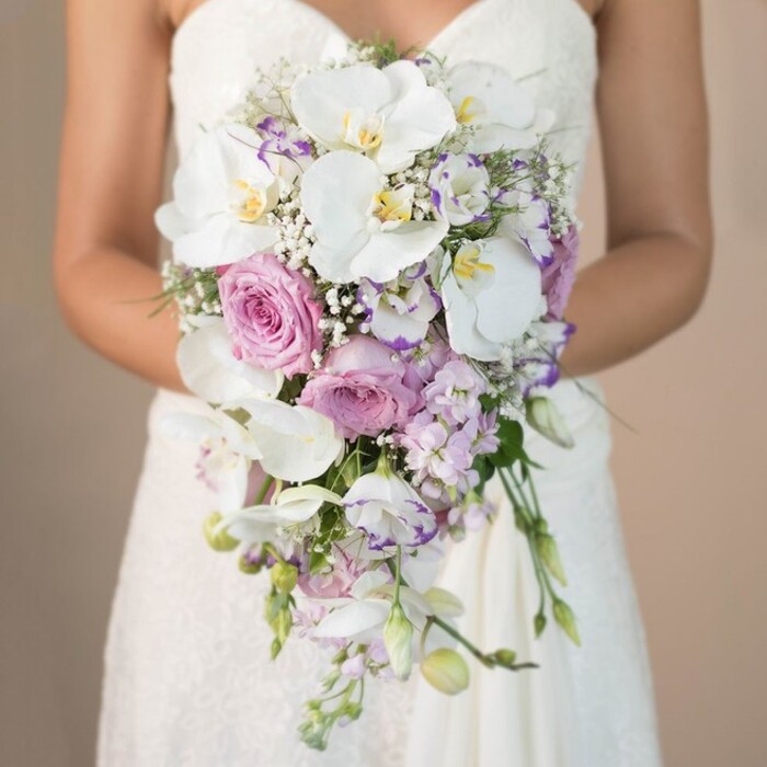 In this radiant, thoughtfully composed photograph, a Stanmore bride is shown from the chest down, standing before a softly blurred, light beige background reminiscent of sun-dappled lawns at Bentley Priory or the elegant interiors of Stanmore's cherished wedding venues. The bride's hands tenderly embrace an extraordinary cascading bridal bouquet, instantly captivating with its harmonious blend of romantic shades. Center stage are flawless, snow-white phalaenopsis orchids, their luxuriously wide petals and golden hearts exuding exotic grace. Around them, soft-pink garden roses bloom in opulent clusters, their tightly curled petals reminiscent of English summer evenings in Stanmore's private gardens. The ensemble is accented by clusters of dainty lavender stock and white lisianthus edged in amethyst-a textural symphony enriched by the cloudy accents of baby's breath threading throughout. Minimal greenery peeks from the lower cascade, contributing a fresh, contemporary feel and subtly referencing Stanmore's leafy surroundings. The bride's strapless gown, rich with lacework seen along the bodice and skirt, peeks through the frame's lower section, its details reflected in the bouquet's artistry. Every element coalesces to evoke an air of sophisticated romance, capturing both the vibrancy and peacefulness of Stanmore. Crafted with care, this bouquet not only acts as the quintessential bridal accessory but also as a celebration of local floral excellence-a reflection of the beauty and charm found within this welcoming North West London community.