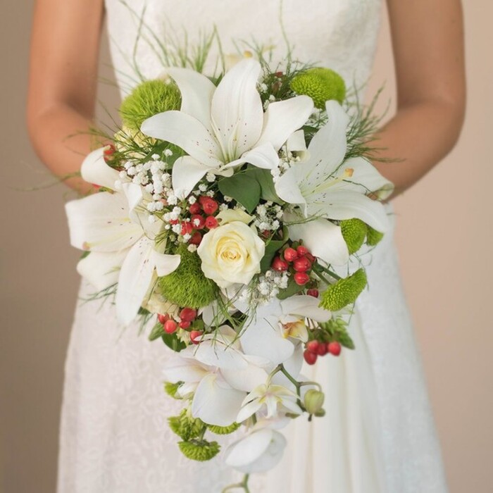 A striking, chest-level bridal portrait set in Stanmore draws the eye to an opulent, cascading bouquet-a testament to the area's botanical richness and the artistry of local florists. The bouquet's prominent feature is its cluster of dramatic white lilies, open and inviting, their petals gently flecked in russet, echoing the natural elegance celebrated in Stanmore's gardens. Centrally, a tightly furling cream rose provides a focal point of romantic anticipation. Encircling the core, vivid lime-green mums burst with energy and freshness, their distinctive pom-pom texture a playful nod to contemporary wedding style. Scattered among these are bright red Hypericum berries, glistening with festive warmth, reminiscent of Stanmore's seasonal markets. Ethereal clouds of baby's breath weave through the arrangement, lending a whisper of lightness that softens the statement blooms. The bouquet's dramatic waterfall effect is created by cascading stems of white Phalaenopsis orchids, their graceful descent evoking the rolling hills and gentle flow of Stanmore's landscape. Wispy, grass-like foliage and thriving green leaves create an abundant, organic frame, bringing the composition to vibrant life. The bride's hands, gently cradling the bouquet against her intricately laced dress, complete the picture of unstudied elegance. With every detail, this bouquet reflects Stanmore's blend of tradition and style-perfectly suited to brides wanting memorable floral artistry for their special day.