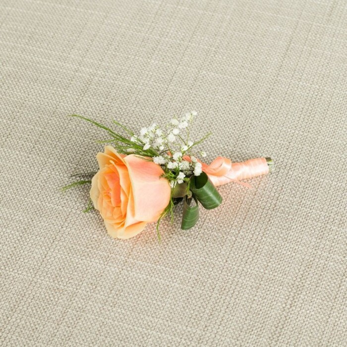 Resting horizontally on a softly textured, oat-beige linen background, this carefully crafted boutonniere centers on a single peach rose, its bloom paused midway between bud and full openness. The petals curl outward in graceful layers, starting with a warm, almost apricot tone deep at the heart and fading into a gentler, creamy peach toward the edges, creating a natural gradient that speaks of freshness and care. Surrounding the upper curve of the rose is a spray of white baby's breath, each tiny floret distinct yet forming a light, airy cluster that softens the composition and introduces a classic, bridal feel. Around the base of the bloom, several strands of slim, bright green foliage stretch out like delicate brushstrokes, while two broader, darker green leaves gently arch and fold around the stem area, visually cradling the rose and adding depth to the colour palette. The gathered stems are tightly bound in a peach satin ribbon that mirrors the flower's tones, its smooth surface wrapped in even spirals and tied off in a small, discreet bow near the lower end. The ribbon catches a hint of the soft, even lighting, giving off a muted sheen that contrasts with the matte texture of the linen beneath and the velvety surface of the petals. The entire piece is photographed in a calm, balanced light, so there are no harsh shadows, only gentle transitions that make every detail easy to trace in the mind. This boutonniere feels ready to be pinned for a wedding in Stanmore, perhaps at a local venue near Stanmore Hill or overlooking nearby green spaces, where such a refined, peach-toned detail would complement a tailored suit and quietly celebrate a day of promises and new beginnings.