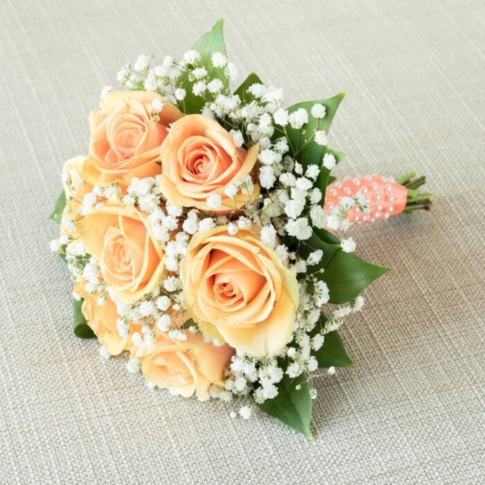 Impeccably composed by local Stanmore florists and presented on soft, oatmeal-toned fabric-perhaps linen or burlap-this bridal bouquet brings together tradition, romance, and local craftsmanship. Its focal point is a grouping of approximately seven perfectly bloomed roses, each one swirling from a tightly furled core outward, petals showcasing a sublime range of peach, apricot, and delicate sunset hues. Interspersed generously among these roses are sprays of dainty white baby's breath, their minuscule blossoms floating like clouds amidst spring meadows, infusing the bouquet with lightness reminiscent of early morning walks through Stanmore Country Park. Flashes of lively green rose leaves emerge here and there, their texture and depth subtly balancing the warmer tones with a flourish that honours Stanmore's reputation for lush, elegant gardens. The stems are expertly bound with a smooth, coral-peach satin ribbon, which spirals with an array of tiny white faux pearls-each one catching the ambient light, evoking memories of pearls and laughter at Stanmore's local wedding gatherings. The bouquet lies slightly angled, revealing its neat, hand-cut stems beneath the crystalline loops of ribbon. Illuminated with gentle, even lighting, every curve and texture is revealed, creating an impression of timeless beauty and heartfelt artistry. Designed for weddings and joyous occasions in the Stanmore community, this bouquet radiates quiet sophistication and enduring love, reflecting the care, precision, and passion of local florists.