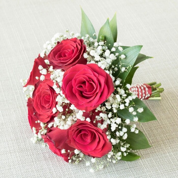 A breathtaking bouquet lies on softly textured beige linen, echoing the graceful sophistication of Stanmore celebrations. Central to the design, five or six plush red roses open to reveal intricate layers of scarlet petals-each one meticulously formed, evoking romance and timeless beauty. Between these radiant blooms, an abundance of pure white baby's breath fills the spaces, its minute blossoms gathered in frothy clusters, delivering a delicate, almost weightless effect that lets the roses shine. Glossy green foliage-likely expertly chosen ruscus-frames each flower, lending a fresh, verdant energy reminiscent of the leafy scenery found in Stanmore's parks. The tightly bound stems are secured with a striking red-and-white striped ribbon, gracefully adorned with a spiral of tiny pearl beads, a decorative touch that hints at bridal elegance and Stanmore's refined sensibilities. Set against a subtle fabric background, the natural daylight brings out the rich, varied tones and the contrasting textures within the bouquet. Whether carried by a bridesmaid at a local wedding, presented at a milestone celebration, or delivered to a loved one in Stanmore, this glorious arrangement is a heartfelt expression of beauty, devotion, and local tradition.