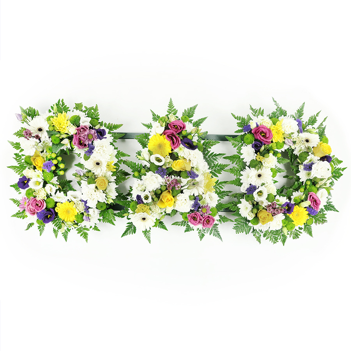In this image, a meticulously crafted DAD floral tribute takes centre stage, set against a bright white background so that every blossom and frond appears crisp and defined. Three robust, block-style letters - D, A, and D - stand side by side, their shapes clearly carved out by a ruffled edging of dark green fern foliage. The fern provides both structure and softness, its natural curves echoing the leafy paths and gardens that characterise much of Stanmore. Within each letter's outline, the florist has woven a dense tapestry of white flowers, primarily plump chrysanthemums and open white gerbera daisies. Their petals, layered and slightly curled, create a smooth, inviting texture that suggests calm, purity, and the stillness of reflection. Dotted throughout the white base, bright yellow gerbera daisies and petite yellow spray roses add cheerful notes, shining like small pockets of light and recalling shared family moments filled with warmth. Sprays of purple lisianthus and rich magenta or deep pink roses provide depth, their saturated tones symbolising enduring love and respect. Tiny clusters of lavender-hued blossoms and bright green button chrysanthemums peek out between the larger blooms, lending fine detail and a gentle sense of movement across the tribute. Thin, dark green bars quietly anchor the three letters along the base, giving the design stability while keeping the focus on the floral artistry and the heartfelt message it conveys. Crafted in a style commonly chosen by families around Stanmore for funerals and memorials, this DAD tribute offers a dignified, deeply personal way to honour a father's life, combining traditional white funeral flowers with softly uplifting colour.