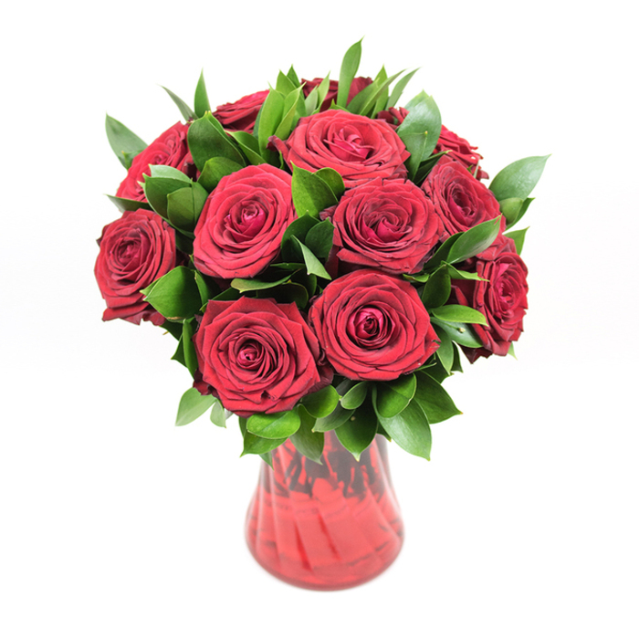 In this image, a classic round bouquet of around twelve deep red roses is carefully composed in a translucent ruby glass vase, set before a stark white background that intensifies the arrangement's rich colours. The roses form a dense, almost perfect dome, each bloom fully open with layers of soft, velvety petals curling outwards in tight spirals. The hue is a deep, romantic crimson, uniform across the bouquet but subtly varied where light grazes the petal edges, creating natural highlights and shadows that suggest depth and movement. Interwoven among the roses is a substantial amount of glossy dark green foliage-likely ruscus or a similar leaf-its pointed, waxy leaves lifting the blooms and framing them, giving the bouquet a full, lush profile rather than a flat, compact look. The foliage also provides a cool counterpoint to the warm red tones, making each rose head stand out more distinctly. The vase beneath is a striking but harmonious partner: its glass is tinted a matching red, with a softly flared opening and an elegant, narrowing curve towards the base. A gentle ribbed or swirled texture along its surface catches reflections, adding rhythm and a sense of crafted detail. Through the translucent glass, darker shadows of stems and gathered greenery can be glimpsed, grounding the bouquet with a natural, almost woodland core. The white background, free of any distractions, transforms the arrangement into a singular visual statement-romantic, polished, and intentional. It feels like the kind of bouquet a Stanmore local might order for a milestone anniversary, perhaps delivered to a home near Bentley Priory or along Marsh Lane, where the timeless message of red roses speaks of enduring love, celebration, and heartfelt devotion.