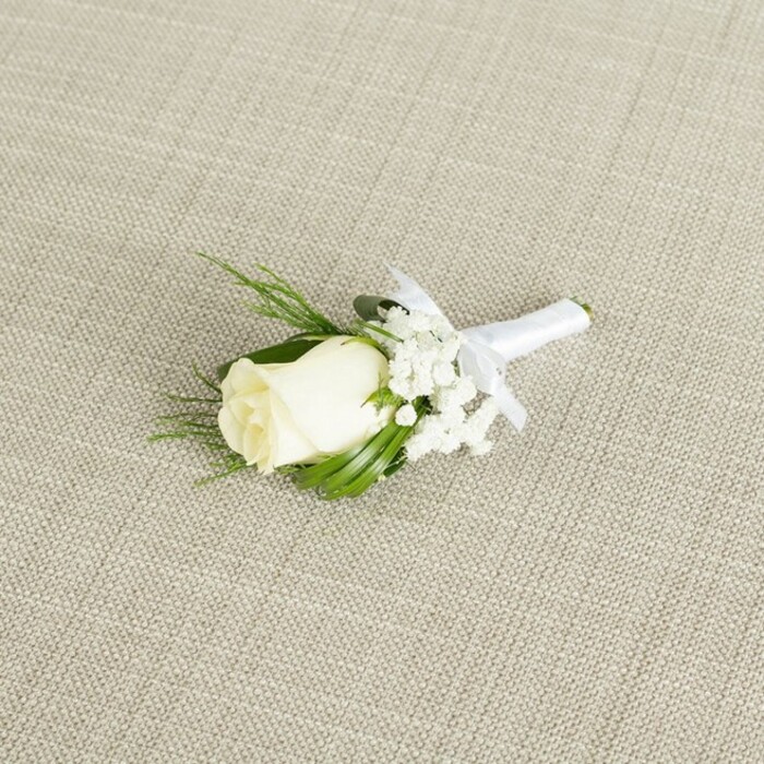 An elegant ivory rose boutonniere is artfully arranged on a softly textured, light beige surface that resembles natural linen, the crosshatched weave lending a quiet, organic feel reminiscent of a carefully dressed wedding table in Stanmore. The boutonniere is positioned at a gentle diagonal, drawing the eye naturally along the line of the stems. At its heart, a single creamy white rosebud sits proudly, the petals smooth, full, and only just beginning to open, giving the impression of freshness and delicate anticipation. Tiny white baby's breath flowers cluster beside and slightly above the rose, their airy, star-like blooms forming a soft, clouded frame around the focal flower. Emerging from behind the rose, several slender deep green leaves and a couple of fine, fern-like sprigs fan out in subtle arcs, adding depth and a natural, garden-grown character that recalls the leafy edges of Bentley Priory Nature Reserve. The stems of the rose, gypsophila, and foliage are drawn neatly together and tightly bound in pristine white satin ribbon, which has a slight sheen that catches the light. Just below the floral head, the ribbon is tied into a small, symmetrical bow, the ends cut cleanly, signalling that this boutonniere is fully finished and ready to be pinned for a wedding or formal event. Soft, diffused light washes over the arrangement from one side, casting gentle shadows that emphasize each curve of the rose and the fine lines of the greenery, while keeping the palette calm and understated. This image captures the type of classic, romantic lapel flower that might be chosen for a groom or best man attending a ceremony in one of Stanmore's churches or function suites, offering a graceful symbol of purity, commitment, and thoughtful preparation on the wedding day.