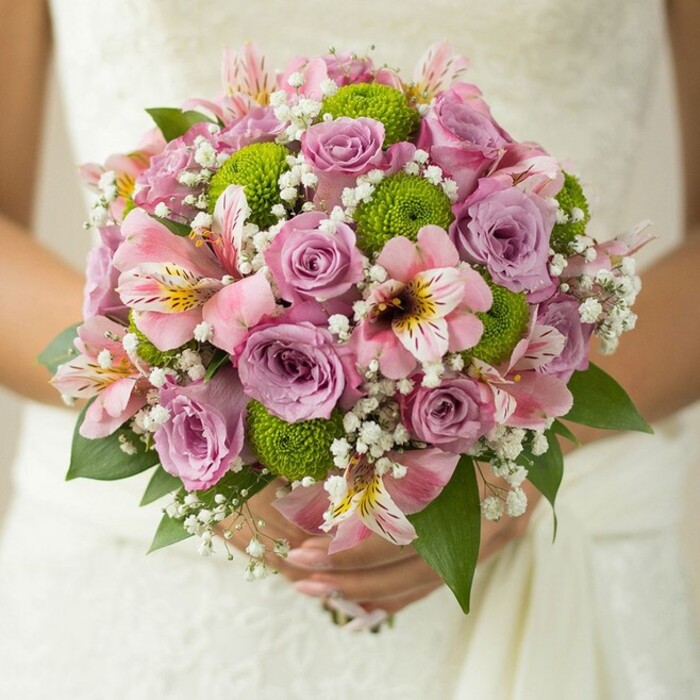 A captivating close-up of a Stanmore bridal bouquet reveals its masterful gathering of blooms, held tenderly by a bride against her flowing, white wedding dress. Central to this lush, hand-tied arrangement are delicate roses in shades of lavender and delicate pink, some petals dewy with the freshness of a Stanmore morning. Lime green button chrysanthemums dot the bouquet, their vivid hues and round forms adding a modern twist to the romantic, pastel heart of the design. Generous pink alstroemeria lilies-marked with signature maroon and gold streaks-peek through, bringing subtle exotic flair and evoking Stanmore's multicultural spirit. Wisps of white gypsophila, airy as the light drifting across the fields at Bernays Gardens, weave between the larger flowers and add an understated softness. Deep green leaves emerge at the base, offering a lush frame that accentuates the bouquet's form. The stems are gently held in hands adorned with a single ring, conveying an atmosphere of joyful anticipation as vows are about to be spoken. Crafted with care by expert Stanmore florists, this bouquet is a testament to floral tradition balanced with cheerful innovation-ideal for spring or summer weddings and for those seeking inspiration grounded in local style.