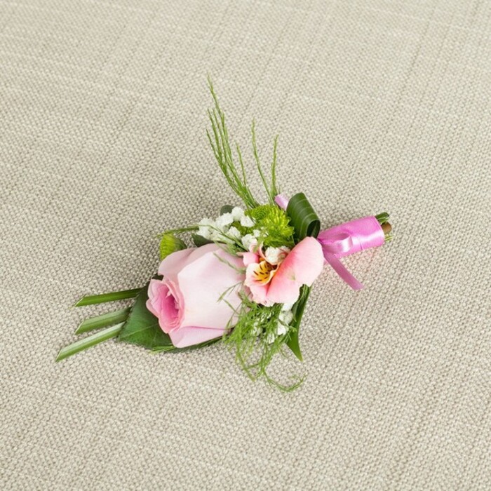 Beautifully crafted and thoughtfully designed by experienced Stanmore florists, this boutonniere presents itself on a softly textured, linen-like fabric in muted neutral tones, reminiscent of timeless interiors found in local Stanmore homes. Its heart is a singular blush pink rose, its petals curled inwards in a perfect spiral, with a deep, warm centre that gently fades to an airy pastel-a symbol of new beginnings. Nestled alongside, a light peach-pink alstroemeria opens wide, revealing a touch of golden yellow and distinct maroon flecks that hint at the intricate detail cherished by both local flower lovers and wedding planners hosting celebrations at Stanmore Country Park. Sprigs of baby's breath, airy and pure, drift among miniature green button mums, offering cloud-like softness and lively accents. The arrangement is framed with feathery, arching fern-like greenery and gracefully broad leaves, some artfully folded to build form and depth. A wide, satin-smooth green leaf wraps elegantly around the bundled stems, finished with a lustrous, fuchsia pink bow that gleams playfully, much like the spirit of a polished Stanmore party. Illuminated by soft daylight, every facet from petals to ribbon glows with freshness and quiet luxury, making this boutonniere, suitable for weddings and formal events, a fine expression of Stanmore's signature floral excellence and understated beauty.