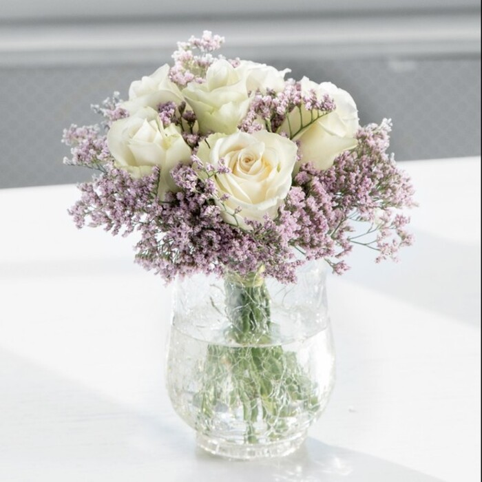 An artfully composed bouquet of creamy white roses and lilac statice stands tall in a clear glass vase, exuding the refined calm so often sought in Stanmore's elegant homes and quiet cul-de-sacs. The arrangement is round and full, with approximately a dozen white roses forming the central cluster. Each rose is at that perfect moment of bloom, where the outer petals have relaxed into soft curves while the inner whorl remains gently folded, promising longevity and grace. Their hue leans towards ivory, with subtle creamy shadows that catch the light and accentuate the petal layers. Interwoven among the roses, wisps of lavender-tinged statice extend outward and upward, each stem branching into countless tiny, star-like blossoms. These miniature flowers introduce a cool, lilac tone and a fine, almost lace-like texture that softens the more structured rose forms, creating the sense of a gentle breeze swirling through a garden. The flowers are anchored in a tall, cylindrical clear glass vase, where neatly trimmed stems are clearly visible, crisscrossing in fresh, vivid green. Clear water fills the base, reflecting flashes of light and hinting at the bouquet's freshness, as if it has just arrived from a local Stanmore florist's workbench. The vase rests on a bright, smooth white surface, possibly a kitchen island or office reception desk, catching soft daylight from a nearby window. The background is intentionally blurred, pale and uncluttered, so the eye lingers on the interplay of cream and lilac, of plush petals and airy sprigs. Subtle whispers of rose fragrance and the faint, dry sweetness of statice come to mind, suggesting a bouquet perfect for gentle condolences, a housewarming near Stanmore Hill, or an understated gesture of affection. The overall feel is modern, serene, and deeply thoughtful, the kind of arrangement that quietly elevates the mood of any room it graces.
