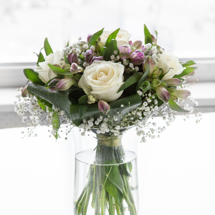 This serene bouquet, captured in soft, luminous light, rests in a clear glass cylindrical vase as if freshly placed on a windowsill in a Stanmore home looking out towards Stanmore Hill. At the core of the arrangement, several creamy white roses bloom with quiet assurance, their petals smooth and velvety, opening in even spirals that feel both timeless and deeply personal. Between these roses, clusters of alstroemeria appear in gentle shades of lavender and purple, tinged subtly with green at their tips, bringing a touch of colour that remains soothing and restrained-perfect for moments of remembrance. Delicate sprays of white baby's breath drift between the larger blooms, filling spaces with a mist-like softness, adding an ethereal, almost weightless quality to the design. Encircling the flowers, rich green foliage creates a lush, polished frame, with broad, glossy leaves and finer, upright stems working together to give structure and depth. The clear glass vase, filled with fresh water, showcases the straight, tidy green stems, a reminder of the natural simplicity and honesty at the heart of the bouquet. In the background, an expanse of bright, diffused white light leaves no visual distraction, allowing every contour of petal and leaf to be seen in the mind's eye. The overall impression is one of composure, grace, and quiet comfort, making this arrangement an ideal choice for sending sympathy to families in Stanmore, Edgware, or along The Broadway. Skillfully created by Flower Delivery Stanmore, it becomes a tender, living symbol of support and remembrance.