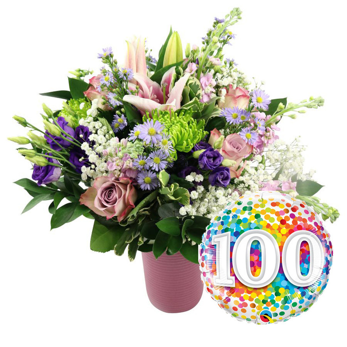 A festive, milestone-themed floral display sits proudly against a crisp white background, creating a striking image that feels right at home in a Stanmore celebration hall or cosy living room. The bouquet is artfully arranged in a modern, cylindrical vase in muted matte pink, its vertical ribbing giving a gentle sense of structure and touchable texture. From this base grows a lavish, dome-shaped arrangement bursting with fresh-cut blooms. Soft pink Oriental lilies stand tall at the centre; some are fully open, with wide, star-shaped petals curling slightly at the tips and long stamens dusted with golden pollen, while others remain as smooth, elongated buds that add height and anticipation. Surrounding them are layers of plush roses in pale blush and deeper mauve or dusty rose tones, their velvety petals forming tight spirals and loose, romantic curls. Interlaced throughout are rich purple lisianthus, their softly ruffled petals bringing depth and drama, and delicate lavender asters with bright yellow centres that lend a cheerful, garden-like charm reminiscent of local Stanmore gardens near Uxbridge Road. Bright lime green button chrysanthemums peek through the pastels, providing vivid contrast and playful, rounded form. Fine sprays of white baby's breath meander through the bouquet like tiny stars, adding lightness and a soft, misty halo. The ensemble is anchored by an array of dark green foliage in varied shapes and sizes, framing the colours and giving the design a lush, full appearance. To the lower right of the vase, angled slightly towards the viewer, a glossy round foil balloon adds a final burst of celebration. Its reflective surface is patterned with multi-coloured confetti dots-reds, oranges, yellows, greens, blues, and purples-surrounding the bold white numerals 100, which are printed large and accented with a faint shadow to give a three-dimensional look. This image captures the feeling of honouring a life or partnership that has spanned a century in Stanmore, making it ideal for representing 100th birthdays, centenary anniversaries, or long-standing community milestones.