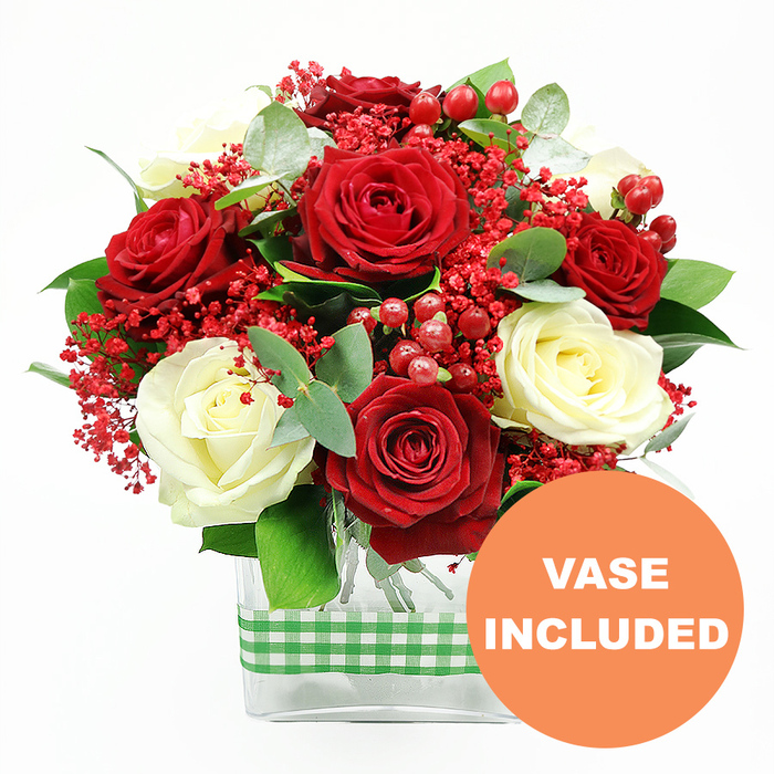 A lush and meticulously crafted bouquet stands out clearly against a plain white background, its colours and textures taking centre stage. The arrangement is dominated by sumptuous red roses and creamy white roses, each bloom at the perfect stage of openness, with petals curled and layered to create a sense of depth and softness. The rich red tones feel passionate and warm, while the creamy roses bring a gentle luminosity, together forming a classic combination that would look perfectly at home in a Stanmore lounge or dining room overlooking local green spaces. Nestled between these larger blooms are delicate sprays of tiny bright red filler flowers, recalling dyed gypsophila or baby's breath, which add a soft, feathery texture and create a subtle haze of colour around the roses. Glossy red hypericum berries are scattered throughout, round and jewel-like, providing sparkles of festive brightness that catch the eye from every angle. Encircling and threading through the arrangement is an assortment of foliage: cool eucalyptus with its silvery-green leaves and richer green foliage that adds structure and contrast, enhancing the sense of freshness and abundance. The stems gather neatly in a clear, rectangular glass vase, whose transparent walls reveal the ordered lines of green stems, emphasising both cleanliness and professional skill. Around the middle of the vase, a band of green and white gingham ribbon is wrapped snugly, introducing a rustic yet cheerful character, almost like a nod to a country-style table set for a family celebration in Stanmore or nearby Harrow Weald. In the lower right portion of the image sits a bright orange circular badge, clearly marked with VASE INCLUDED in bold white letters, showing that this is a complete, ready-to-display gift, ideal for anniversaries, romantic surprises, or heartfelt same-day deliveries across the local Stanmore community.