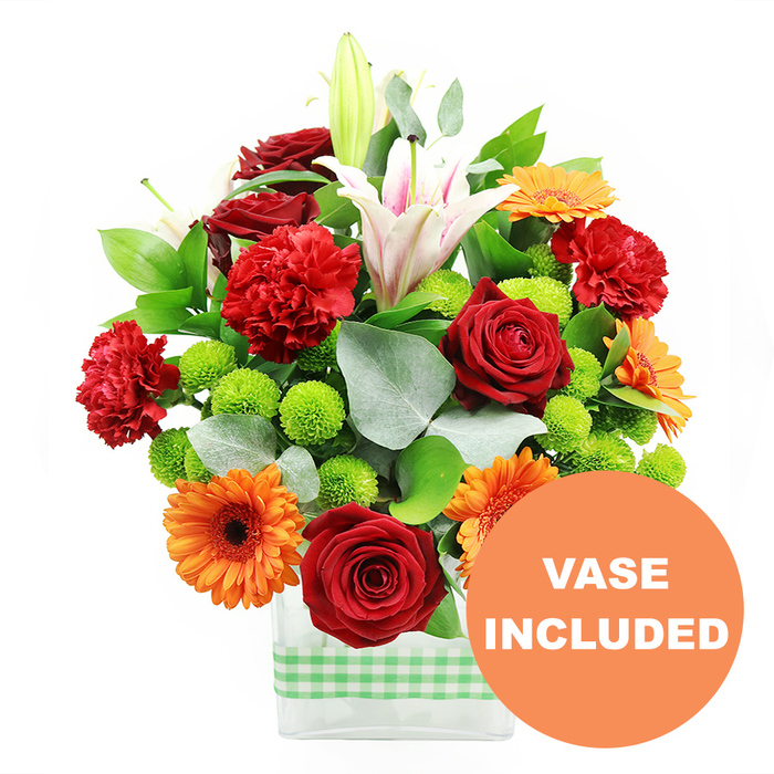 This photograph features a vibrant mixed bouquet arranged with the care and precision of an experienced florist, presented in a contemporary rectangular glass vase. Around the lower half of the vase runs a pale green and white gingham ribbon, adding an approachable, homely accent that would suit a kitchen window or dining table in Stanmore. The bouquet itself is full and rounded, overflowing with rich, saturated colour. Deep crimson red roses, their petals densely furled and velvety, anchor the design, conveying a sense of heartfelt affection. Beside them, red and red-orange carnations introduce a soft, frilled texture that gives the arrangement a plush, cushion-like effect. Scattered among these, bright orange gerbera daisies with dark, almost chocolate centres bring a playful brightness; their flat, radiating petals create bold circular bursts that catch the eye the way a stand-out shopfront might along Church Road. Clusters of tight, spherical lime-green button chrysanthemums nestle between the larger blooms, their fresh, zesty hue adding a lively contrast and helping to link the flowers visually to the ribbon around the vase. Poised above and through the mix, several white Oriental lilies add height and a graceful, sculptural dimension; some are fully open, revealing delicate pink veining and pollen-tipped stamens, while others are still in bud, promising movement and change in the days after delivery. A generous collar of glossy deep green foliage, interlaced with cool, silver-green eucalyptus leaves, frames the colourful flowers, providing depth, balance, and a subtle sense of woodland calm. The clean white background removes all distraction, focusing attention entirely on the interplay of colours and shapes. In the bottom right of the image, a bold orange circular emblem with white capital letters clearly states VASE INCLUDED, signalling that this arrangement arrives as a complete, ready-to-display floral gift for celebrations and thank-yous around Stanmore.