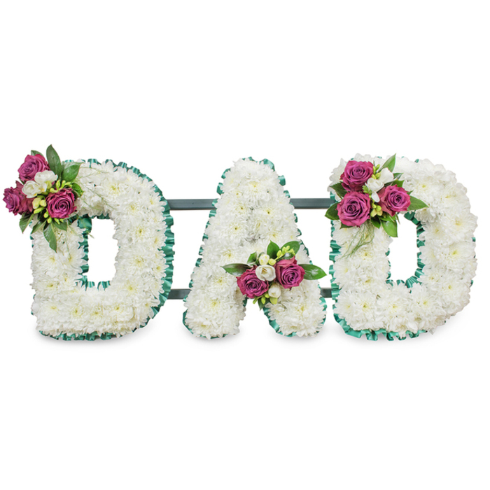This elegant sympathy piece spells out the letters DAD in capital form, each letter lovingly constructed from a thick layer of pristine white chrysanthemums that give a velvety, tactile surface against a stark white background. The tiny, cushiony florets nestle together to form bold, readable shapes whose soft petals diffuse light and suggest a sense of peaceful clarity. A vibrant emerald-green ribbon-pleated or gently ruffled-traces the perimeters and inner openings of each letter, creating a clear, solemn outline and a refined visual contrast to the white blooms. A discreet dark green or grey bar runs behind the letters, implying the easel or support that will carry this design to a funeral, memorial service or graveside.
Thoughtful accent clusters of deep pink to magenta roses, paired with delicate white freesia and full, glossy foliage, are positioned to balance the composition: the first D bears its cluster at the upper left curve, the A is graced with a group in its lower V, and the final D reflects the first with a cluster on its upper right curve. The roses lend a plush, tactile warmth while freesias add a soft citrus-honey scent; the greenery brings contrast and structure. I can imagine this piece positioned in the quiet light of a chapel near Stanmore Broadway or set beside a headstone close to Stanmore tube station, offering a clear, heartfelt expression of love, respect and farewell.