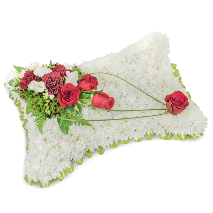 A dignified rectangular pillow of sympathy flowers sits against a neutral backdrop, its entire surface meticulously formed from hundreds of small white chrysanthemum blooms packed so closely they read as a single, velvety plane. The craftsmanship is evident in the gentle rise of the cushion and in the way the white petals catch soft light, creating a calm, luminous field that evokes rest and purity. Around the perimeter a whisper of pale green ribbon or slim foliage edges the composition, providing a neat, refreshing frame. At the upper left an elaborate floral cluster introduces deep colour and texture: several sumptuous red roses, some tightly furled and others in fuller bloom, are paired with frilly burgundy carnations and spotted with tiny white freesia-like sprays that add delicate sparkle. Lush greens - including slender fern fronds and denser leaves - lift and cradle the blooms, giving depth and a reassuring verdant backdrop. From this corner a trio of graceful green lines, likely bear-grass or long decorative foliage, arcs diagonally across the white mattress toward the lower right, their elegant sweep linking the composition and guiding attention to a single, immaculate red rose positioned there. That solitary bloom, gently resting on the chrysanthemum surface, feels like a final, intimate gesture of love. The overall impression is one of composed sorrow and tender remembrance, suitable for memorial services at local venues near Stanmore High Street, St. John's Church or the tranquil green spaces around Stanmore Common, where families often place tributes in honour of a loved one.