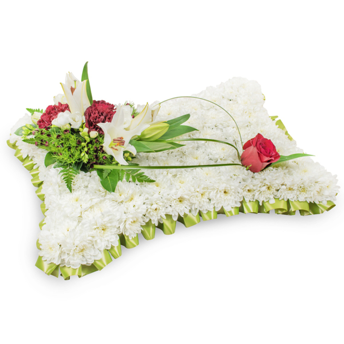 A classic rectangular funeral pillow composed entirely of small, pristine white chrysanthemums, each bloom densely set to create a smooth, soft-textured surface that suggests quiet rest. The edge is trimmed in a pleated pale lime-green satin ribbon, folded with exacting care so the border resembles a neat, gentle frill. Nestled at the upper left is a composed cluster of white Oriental lilies - two blossoms opened to reveal their yellow stamens and a single slender bud - paired with deep burgundy spray carnations whose rich tones and ruffled petals form a warm counterpoint to the ivory cushion. Mixed foliage, including feathery fern fronds and larger glossy leaves, provides a lush, green backdrop and subtle variations in texture. From this cluster a slim, arching loop of bright green salal leaf or decorative wire stretches toward the right-hand side of the pillow, where it naturally settles at the tip beside one deep red rosebud, poised as a solitary emblem of enduring affection. The velvet-like rosebud's colour and form draw the eye across the composition, completing a visual journey from left to right. Lit softly against a white studio background, the arrangement conveys purity, dignity and heartfelt sympathy. Crafted with traditional funeral etiquette in mind, this floral cushion is a considered expression of remembrance for a memorial service or wake; Flower Delivery Stanmore can prepare similar tributes for local deliveries around Stanmore High Street, Stanmore Station and Stanmore Common with attentive, neighbourhood care.