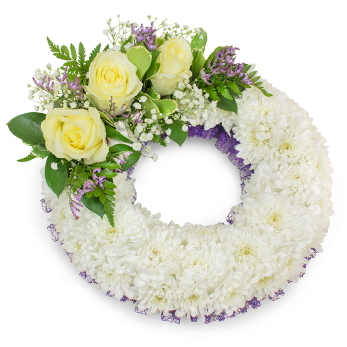 A serene, formally composed memorial wreath set against a stark white backdrop, its principal mass formed from hundreds of pristine white chrysanthemum blooms arranged so densely that the surface appears like a soft, woven cushion. The chrysanthemums' layered petals create a full, tactile texture - cool to the eye and soothing in its uniformity - while a subtle band of purple netting or ribbon slips through the inner and outer margins, lending a refined hint of colour and dimension. Nestled on the upper-left arc is a considered cluster of three creamy yellow roses, their petals unfolding in gentle spirals that radiate warmth and friendship. These roses are accompanied by delicate sprigs of white gypsophila and small, papery lavender statice which offer delicate points of contrast and a faint herbal scent. The greenery around the cluster - broad, shiny leaves contrasted with finer, fern-like fronds - frames the flowers and enhances the wreath's visual balance. The design preserves a clear central aperture, a classic symbol of eternal remembrance, and the overall effect is one of calm dignity suited to a funeral, wake, or memorial service. As a local Stanmore arrangement familiar to florists delivering to nearby Harrow Weald or Pinner, this wreath communicates compassion and respect: understated, expertly crafted, and meant to soothe grieving families with its quiet beauty and thoughtful detail.