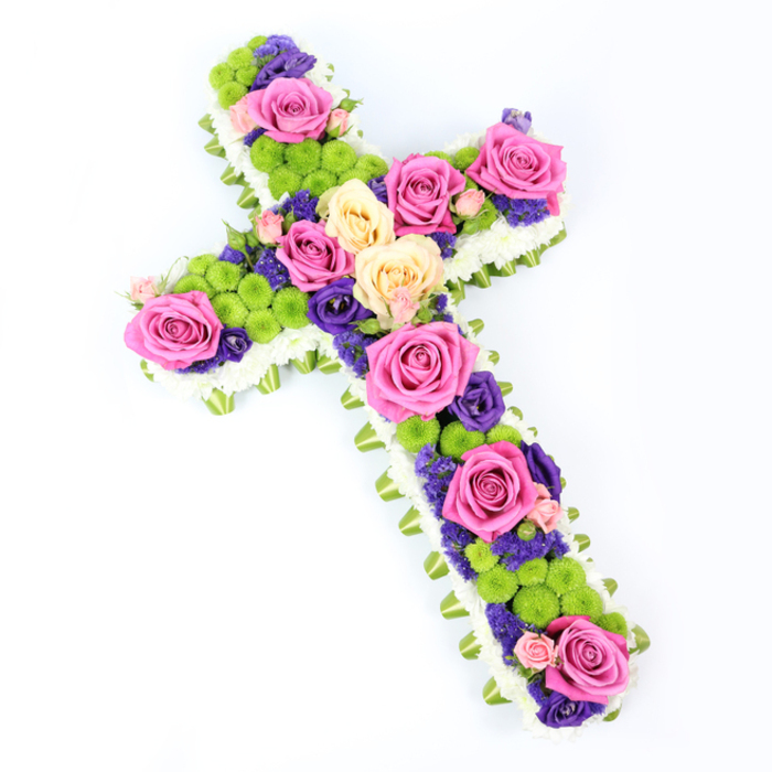 Photographed on a clean, high-key white background, this floral cross arrangement appears luminous, its colours vividly defined under bright, even lighting. The cross itself is slightly tilted on the page, running from the upper left to the lower right, which gives the composition a natural, almost graceful movement. Around the outer edge, a dense line of white spray chrysanthemums traces the full shape, each tiny daisy-like floret nestled closely against the next to create a soft, cushiony frame. Just beneath this delicate border, a narrow band of light-green ribbon or neatly folded foliage peeks out, adding a gentle halo of green that emphasises the cross's silhouette and reflects the lush greenery seen in Stanmore's parks and gardens. Inside this border, the central design is a lush tapestry of roses and chrysanthemums. Large hot-pink roses and softer, creamy pale yellow roses are distributed evenly across the arms and along the length of the cross, their petals fully unfurled, catching the light and providing focal moments of beauty. Punctuating these roses, small clusters of lime-green button chrysanthemums create a lively texture, their rounded, almost spherical heads forming bright, cheerful notes within the more formal tribute. Threaded between and around the larger blooms are smaller deep purple and violet flowers, most likely lisianthus or statice, which bring a rich, jewel-toned contrast and add visual depth, much like shadows in a painting. The result is a funeral flower cross that feels both reverent and uplifting, an ideal tribute for services at local Stanmore churches or crematoria, conveying remembrance, faith, and a quiet sense of hope.