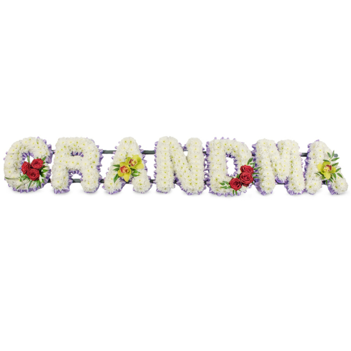 This image shows a lovingly designed GRANDMA funeral tribute, the seven capital letters set side by side and photographed front-on against a plain white background. The clean backdrop throws all attention onto the flowers, making the tribute feel bright and contemplative. Each letter is generously covered with small white chrysanthemum blooms, meticulously arranged so that they form a dense, velvety layer with no stems or gaps showing. The effect is of a soft, white cushion, inviting thoughts of peace and rest. Just beneath the main layer of white, a thin rim of pale lavender or soft purple flowers runs along the base and edges, creating a delicate outline that gently emphasises the shape of each letter without disturbing the overall calm. The design has been given extra character through a few well-placed accent flowers. On the lower curve of the G, three deep red, half-open roses are nestled into a bed of dark green foliage, their warm hue symbolising devotion and gratitude. The first A features two yellow-green orchids gently placed across its bar, their speckled, reddish throats adding fine detail and a hint of luxury. The R, N, and D in the centre of the word remain entirely white and lavender, keeping the heart of the tribute serene. The M repeats the rose motif, with three more red blooms grouped on its lower right stroke, and the final A closes the word with another pair of chartreuse orchids. A discreet dark green or black metal frame supports all the letters from behind, prepared for display at a church, cemetery, or crematorium in Stanmore, such as those near Stanmore Country Park. The overall feeling is one of love expressed through calm, refined floristry.