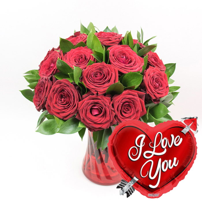 This image presents a richly coloured romantic arrangement: an opulent gathering of deep red roses paired with a gleaming heart-shaped balloon, set cleanly against a solid white background. At the centre, around a dozen or slightly more roses are tightly massed together in a classic, rounded bouquet. Each bloom is a deep crimson, the petals thick and velvety, some curling inward like whispered secrets, others opening outward to show layers of soft, overlapping petals. Dark emerald-green foliage surrounds and supports the roses, its leaves smooth and full, lending the bouquet a fresh, garden-grown feel reminiscent of weekend walks through the green spaces near Stanmore Country Park. The flowers rise from a clear ruby-red glass vase, which features delicate vertical flutes or ridges running up its sides. These ridges catch the light and create gentle reflections, while the tinted glass subtly colours the water inside, drawing attention to the crossed stems that anchor the arrangement. In the lower right portion of the image, partly overlapping the vase and bouquet, is a bright red heart-shaped foil balloon. Highly polished and reflective, the balloon appears almost mirror-like, its metallic surface dotted with soft highlights. Across the centre of the heart, the words I Love You are written in sweeping white cursive letters that curve gracefully with the balloon's shape. A silver Cupid's arrow is depicted as if piercing through the heart on a diagonal, a classic emblem of love struck suddenly and deeply. The uncluttered white studio background accentuates every nuance of red and green, making this arrangement look like an ideal gift for Valentine's Day, an engagement celebration, or a surprise romantic delivery anywhere across Stanmore.