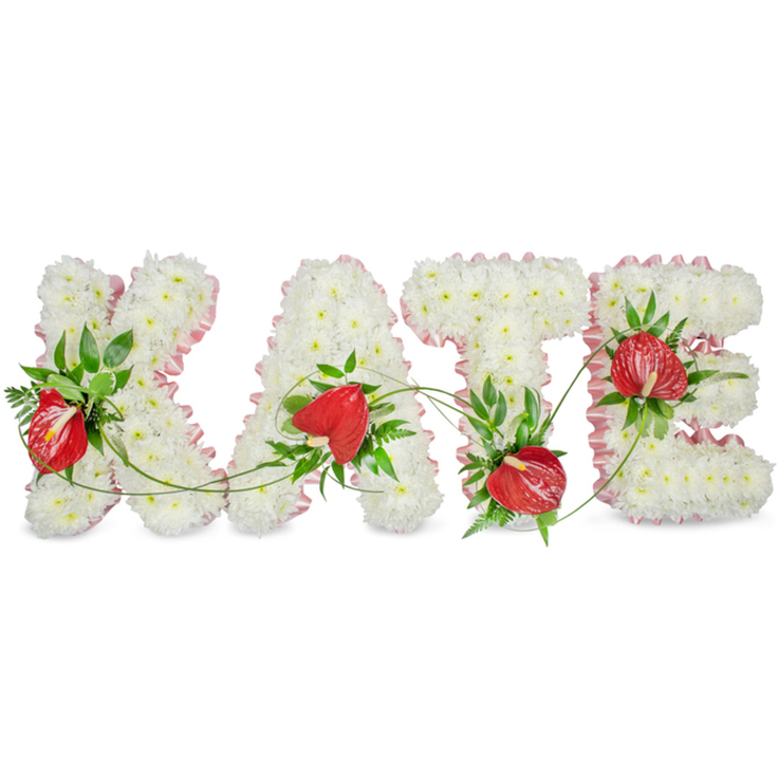 A refined and dignified floral name tribute reads KATE in bold, sculpted capital letters, each letter meticulously covered with a dense blanket of small white spray chrysanthemums that create a seamless, soft texture evoking purity and repose. Along the outer edge of each letter a pale pink ribbon or fabric trim has been applied with care, its subtle pleating introducing a feminine, comforting accent against the immaculate white blooms. Interspersed through the chrysanthemum base are glossy red anthurium blooms, selected for their dramatic, heart-shaped spathes and prominent spadices that provide a striking visual counterpoint and focal warmth. The anthuriums sit among rich, dark green foliage - broad leaves, glossy ruscus and delicate fern fronds - which are arranged to frame and lift the red accents while adding natural depth and sculptural line. Fine green vines trail and bridge the negative space between the letters, lending unity and a gentle, living motion to the composition. A faint shadow beneath the letters gives them a subtle three-dimensional lift against the bright white background, making the forms read clearly from a distance. The overall impression is one of composed elegance and heartfelt remembrance, suitable for services at local venues in Stanmore, near Stanmore Station or the quiet avenues around Stanmore Common. As a bespoke funeral tribute, this arrangement communicates respect, enduring affection and the quiet craft of experienced local florists who understand what families in the area seek at times of loss.