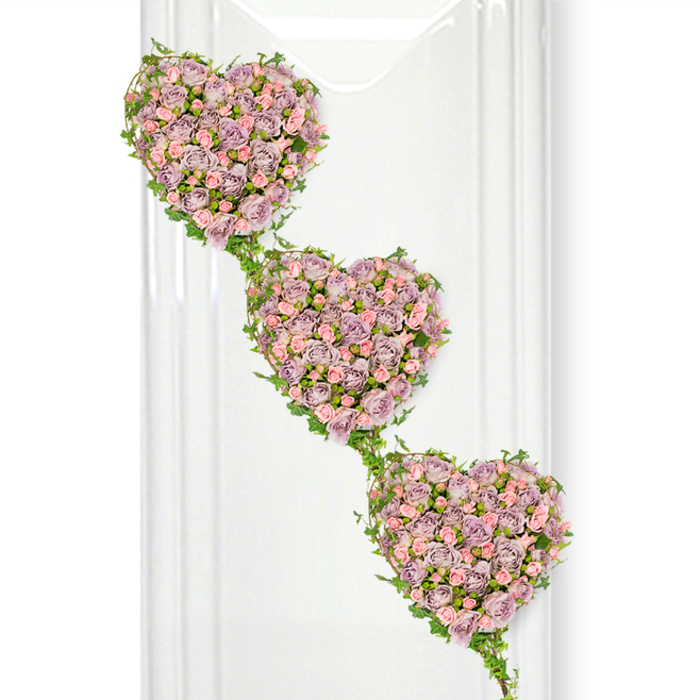 A delicate cascade of three rose-filled hearts forms the centrepiece of this dignified funeral tribute, floating against a pure white background that lends the design a sense of calm and space. The hearts are aligned on a soft diagonal, stepping gently from the upper left down toward the lower right, each one intimately packed with miniature roses in subtle shades of dusty lavender, muted mauve, and tender pastel pink. The roses sit so closely together that they form a smooth, cushioned surface, while the interplay of buds and open blooms adds texture and a feeling of quiet life. The colour palette feels soothing and nostalgic, like a treasured memory recalled at a peaceful moment. Threading their way around the edges of the hearts and drifting between them are fine strands of green ivy. The small, glossy leaves follow the heart outlines and then stretch out to join one heart to the next, creating a natural, flowing link that echoes themes of continuity, connection, and an unbroken chain of love. Behind the arrangement, the bright white setting is enlivened by subtle vertical reflections that hint at a contemporary, light-filled room, perhaps similar to a chapel or reception space in Stanmore where families gather after a service. There is no clutter, only the gentle contrast between the pastel roses and the vivid greenery. This is the kind of thoughtfully composed tribute Flower Delivery Stanmore might create for a funeral, capturing the emotions of tenderness, gratitude, and enduring affection that families wish to express when saying goodbye.