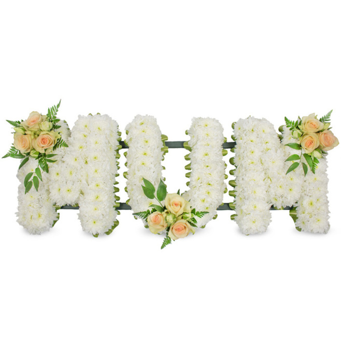 A refined sympathy arrangement in the photograph spells the word MOM in large, block capital letters, stretching horizontally across the frame. Each letter is meticulously built from hundreds of small white flowers, almost certainly spray chrysanthemums or similar blooms, carefully inserted so closely together that they create a uniform, snowy-white surface. The M, O, and M share the same height and width, sitting steadily on an unseen base, with just the faintest hint of a dark green support visible at the sides and in the gaps between some blooms, giving definition to the lettering. Resting atop this field of white are three accents of colour that soften and personalise the design. Small clusters of peach and apricot spray roses are positioned with florist's precision: at the upper left of the first M, at the lower centre of the O, and at the upper right of the last M. The roses display gentle, rounded petals that range in tone from blush to a slightly deeper, warm peach, and they are accompanied by fresh green foliage, including airy ferns and more solid leaves. These touches of greenery create shadows and layers, giving the tribute a sense of depth and life rather than appearing flat. The clean white background allows the eye to focus entirely on the word and its soft colours, making it easy to imagine this arrangement placed at a Stanmore crematorium chapel or carried carefully from a local florist like Flower Delivery Stanmore to a family home. The design radiates quiet respect, comfort, and heartfelt love for a mother, offering a visual message of remembrance that is humble yet deeply moving.