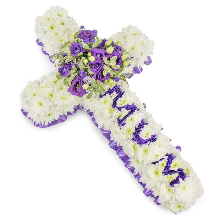 An elegant, large-scale floral cross presented against a crisp white field, composed chiefly of compact, creamy-white chrysanthemum blooms whose cumulative surface creates a serene, cushiony texture. The entire perimeter is traced with a deep purple satin ribbon, its ruffled edge lending a formal, respectful frame to the silhouette. Along the vertical axis, the word MUM is rendered in distinct purple flower clusters - small, round heads of eustoma or spray chrysanthemums - each letter clearly modelled to stand out against the white foundation as a personal dedication. At the central junction, a rich, multi-tonal cluster becomes the focal point: velvety deep violet roses paired with lighter lavender lisianthus (eustoma), layered amid variegated green leaves whose pale margins catch the light and create visual depth. Speckled green berries and slender foliage stems weave through the cluster, introducing subtle texture and a sense of natural calm. The overall colour story - pure white balanced with a spectrum of purples from soft lavender to deep violet - communicates dignity, peace, and sorrow in equal measure. Photographed in even, diffused light, the arrangement's textures and colours are clear and tactile. As a local Stanmore floral designer I would suggest this cross for a funeral service at a local church or a graveside near Stanmore Common, where its quiet fragrance, composed lines, and meticulous craftsmanship will offer a heartfelt tribute and a consoling presence for mourners.