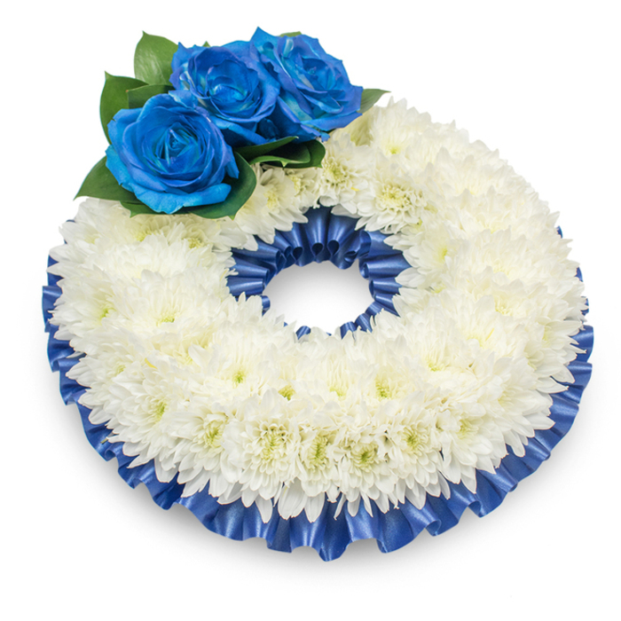 A refined circular wreath presented against a bright white backdrop, photographed slightly from above so its dimensional form and careful detailing are clearly visible. The principal element is a dense ring of immaculate white chrysanthemums: layered, feathery petals arranged into a slightly raised band that suggests both softness and ceremonial formality. Accenting the wreath at the top-left are three intense blue roses-deep cobalt to sapphire-each blossom showing the velvety spiral of petals that modern dye processes produce without losing natural shape; these are nestled into clusters of rich, dark green foliage which both anchor and lift the blue blooms against the white field. A matching pleated satin ribbon trims the wreath's outer and inner edges, its subtle sheen and ripple giving definition and a composed finish to the arrangement. The gentle shadow beneath the wreath provides depth and grounds the piece on the white plane, while the overall colour story-pure white tempered by a solemn, cooling blue-speaks of remembrance, respect and calm. As a local florist familiar with memorials in and around Stanmore High Street, Canons Park and the churches near Harrow, I choose materials and proportions so the wreath conveys dignity from any vantage: the chrysanthemums' soft texture soothes visually, the roses' velvet richness lends a personal touch, and the ribbon frames the sentiment. This arrangement is suited to funeral services, wakes, or floral tributes where understated grace and heartfelt condolence are required.