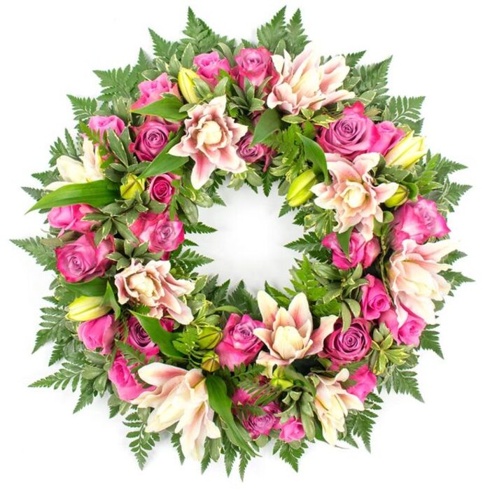 This image features an elegant circular wreath, clearly intended as a funeral or sympathy tribute, photographed against a bright, uncluttered white background that enhances its every detail. The wreath forms a complete, continuous ring, with no bare spaces, just a dense tapestry of flowers and foliage. The primary blooms are roses in shades of vibrant fuchsia, intense magenta, and softer pink, each flower boasting gracefully layered petals that range from tightly closed buds to lush, fully opened blossoms. Interspersed with the roses are generous double lilies, their multi-layered petals opening in tones of creamy white, blush pink, and pale peach, with a few petals tinged with slightly deeper pink toward the tips. Here and there, upright lime-green lily buds emerge, adding a sense of freshness and hope to the composition. All of the blooms are nestled into a bed of luxuriant greenery: feathery fern fronds create a soft, textural edge, while broader, glossy leaves in deep, rich greens fill the inner spaces and provide a strong visual foundation. The combination of verdant foliage and warm-toned flowers creates a balanced, harmonious look that feels appropriate for dignified services in and around Stanmore, whether at a local chapel, crematorium, or cemetery. The overall effect is one of calm elegance and heartfelt sentiment, as if the wreath were crafted by a Stanmore florist attuned to the community's traditions and the need for gentle, respectful expressions of sympathy during times of loss.