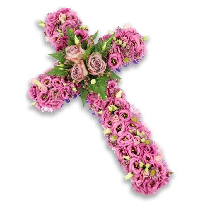 An ornate floral cross tribute is shown centered on a brilliant white background, the slight drop shadow gently lifting the design and emphasizing its three-dimensional form. The entire cross is intricately covered in flowers, dominated by soft pink, mauve, and muted purple hues that blend together in a soothing, harmonious palette. At the point where the vertical and horizontal arms meet, three generously open roses in dusty-rose and light mauve tones create a strong focal cluster, their velvety petals unfurling in layered spirals. Surrounding these central roses is a collar of lush greenery, including airy fern fronds and smaller, deeper green leaves that provide contrast and echo the calming greens seen around Stanmore's quiet residential streets and nearby woodlands. From this centre, the arms of the cross stretch out, entirely cloaked in trumpet-shaped lisianthus blooms. Their ruffled petals overlap like soft fabric, moving through shades from bright, cheerful pink to soft blush and richer mauve, with some flowers featuring darker centres that gently deepen the overall effect. Scattered through the lisianthus are tiny clusters of pink and lilac filler flowers and a sprinkling of pale cream blossoms with delicate yellow hearts, adding fine detail. Small, light green buds emerge here and there, suggesting continuity and hope. Along the very edges of the cross, slender strips or hints of dark-purple ribbon or structural backing peek out, tracing the outline and revealing the careful craftsmanship behind the design. The arrangement carries a mood of calm reverence and heartfelt remembrance, perfectly suited for funeral or memorial services held in and around Stanmore. It offers a graceful way to honour a loved one's faith and memory, combining soft colour, abundant texture, and thoughtful composition.
