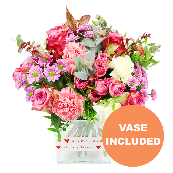 A generously full bouquet stands tall in a clear glass cube vase, dominating the image against a clean white background that makes the colours appear vivid and fresh. The composition feels carefully balanced, as though created by an experienced Stanmore florist for same-day delivery across the local area. At the centre, several large roses in harmonising shades of pink draw the eye: some blooms are a deep, saturated fuchsia with velvety petals, while others are softer blush, opening in gentle spirals that suggest tenderness and affection. Encircling these are lush carnations in peach-pink, coral, and creamy white, their frilled, layered petals adding a rich texture and fullness to the design. Between the larger blossoms, smaller daisy-like chrysanthemums in lavender and muted purple peek through, their bright lime-green centres providing a striking, cheerful accent that stops the palette from feeling too heavy. A generous collar of foliage frames the flowers, including rounded, silvery-green eucalyptus leaves, darker glossy greenery, and sprigs tipped with reddish-brown for a quiet touch of warmth. The stems can be seen clearly through the smooth sides of the square vase, giving a sense of order and freshness, as if the bouquet were just placed on a windowsill overlooking Stanmore Country Park. Around the base of the vase, a white band is neatly wrapped, decorated with repeating red heart motifs and the loving phrase all my love in a handwritten-style red script, turning the vase into part of the sentiment. In the bottom right of the image, an orange circular emblem with bold white capital letters announces VASE INCLUDED, making it clear that this is a complete, ready-to-display gift. The overall impression is of a bright, affectionate arrangement that would suit romantic gestures, family celebrations, or thoughtful thank-you gifts.