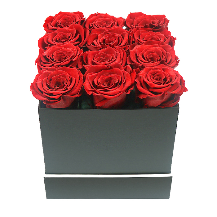 A refined bouquet of twelve preserved ruby red roses is displayed in a compact square black box, captured in a close-up, eye-level shot that emphasises both craftsmanship and elegance, much as you might expect from a dedicated Stanmore florist. The roses are evenly spaced in a 3x4 grid, each bloom perfectly open and uniform in size, their velvety petals forming tight swirls that build from the outer edges towards a deep, shadowed centre. The colour of the petals is a sumptuous crimson, with gentle variations of darker red hidden within the folds, adding richness and dimension. The flower heads are packed so closely that they form an almost seamless cushion of red, just cresting the top edge of the box to create a sense of fullness and luxury. The container is a modern, matte black square with smooth, crisp sides and defined corners, its understated design allowing the bold colour of the roses to dominate. Around the lower section of the box runs a fine white stripe that reads like a minimalist belt, bringing a hint of graphic contrast without overwhelming the design. The entire arrangement is set against a flawlessly white background, free of any other detail, so the eye remains focused solely on the interplay of red, black, and white. Soft, diffused lighting kisses the petals and box edges, casting gentle shadows that suggest the texture and depth of the arrangement. The overall mood is one of lasting romance, contemporary style, and thoughtful gifting, ideal for delivery to homes near Stanmore Golf Club, quiet avenues around Belmont Circle, or the residential streets that wind towards Harrow Weald.